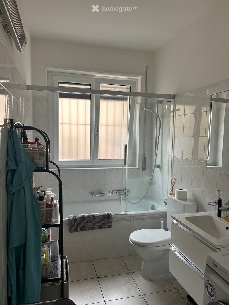 Modern bathroom with a bathtub, toilet, sink, and shower area. Features a large window with frosted glass, marble tiles, and a towel on the edge of the bathtub. A robe and various toiletries are placed on a rack near the window.