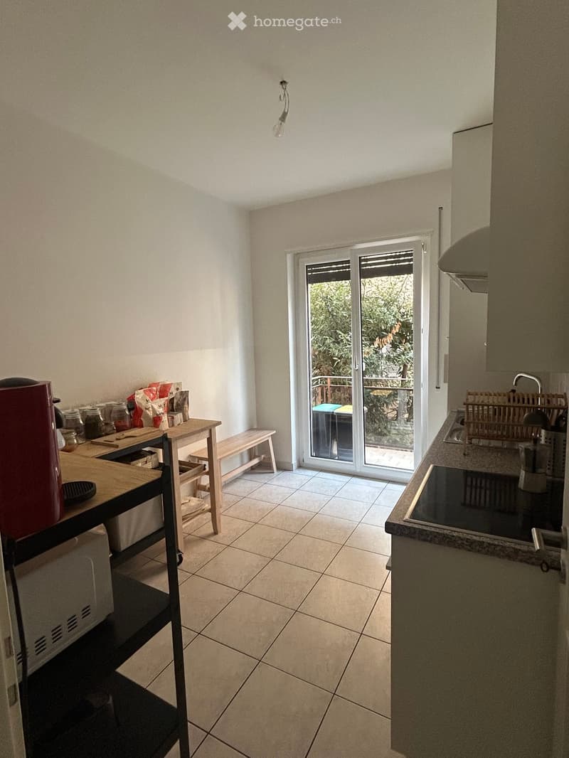 Bright kitchen with tiled floor, white walls, and a sliding glass door leading to a balcony. Includes a wooden table with a bench, coffee maker, microwave, and sink with a faucet.