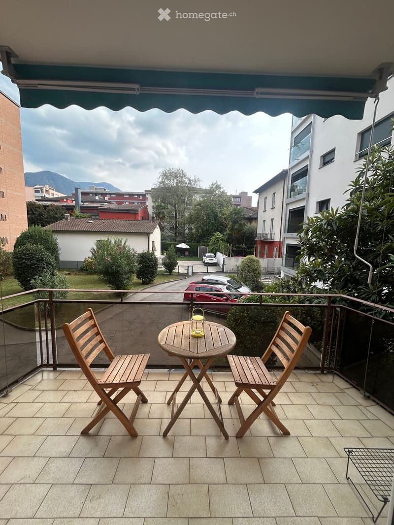 Balcony with a small round wooden table and two wooden folding chairs, overlooking a residential area with parked cars and greenery.