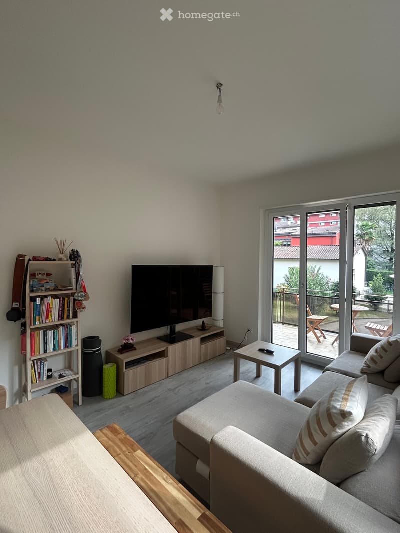 Modern living room with a grey sectional sofa, coffee table, TV stand, bookshelf, and a sliding glass door leading to a balcony.