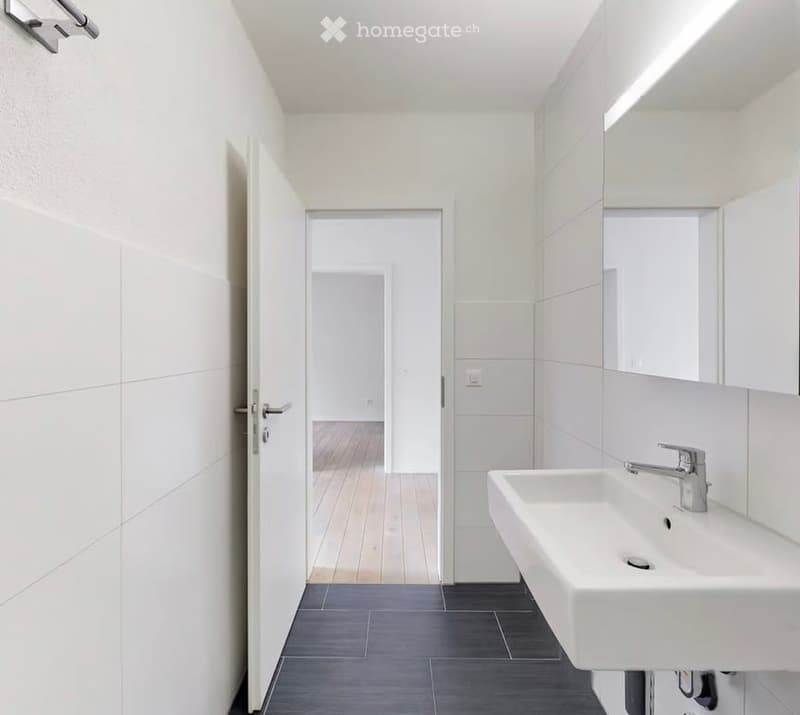 Modern bathroom with white tiles, dark grey floor, white sink, chrome faucet, and large mirror.