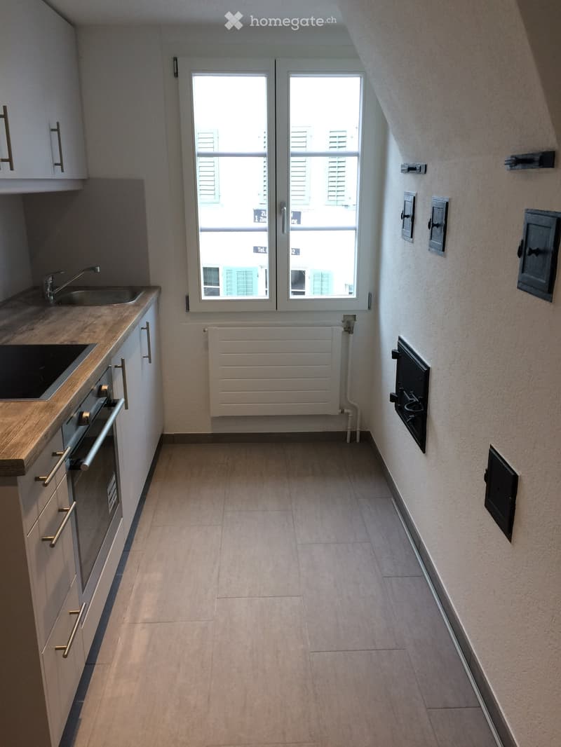 Empty hallway with tiled floor, white walls, and a window at the end. Kitchen area visible on the left with cabinets, sink, and stove.