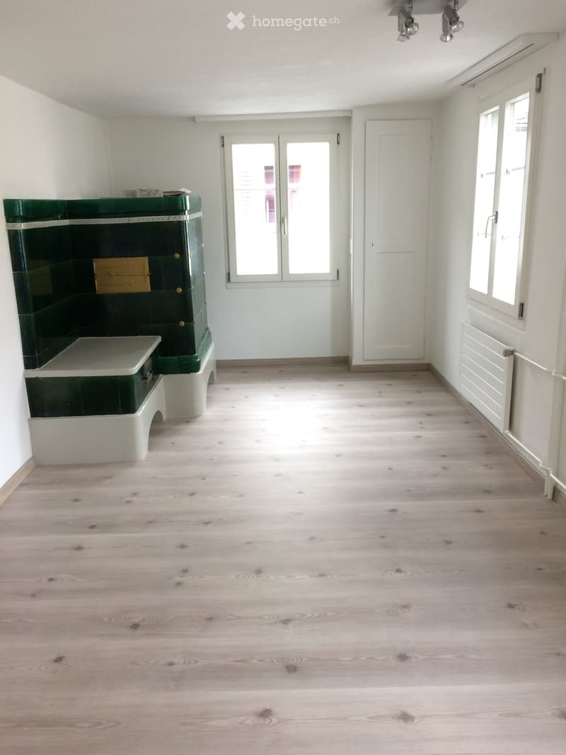 Empty room with light wooden flooring, white walls, and a green fireplace on the left side. Two doors and windows are visible.