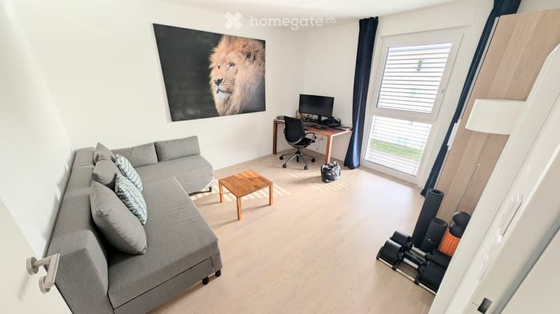 Modern living room with grey sectional sofa, wooden coffee table, desk with computer, and large window with blinds.