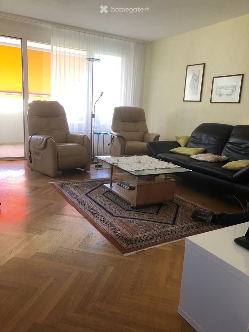 Living room with wooden flooring, large windows, a black leather sofa, two armchairs, a glass coffee table, and a floor lamp.