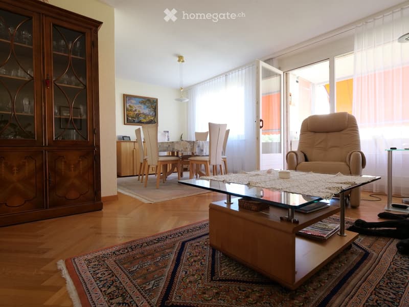 Bright living room with wooden floors, large windows, a glass coffee table, a beige sofa, a dining area with chairs and a table, and a wooden cabinet with glass doors.