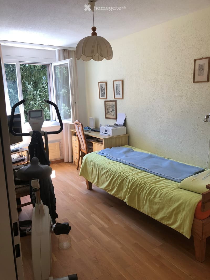 Well-lit bedroom with wooden flooring, a single bed with green and blue bedding, a desk with a chair, an exercise bike, and framed pictures on the wall.
