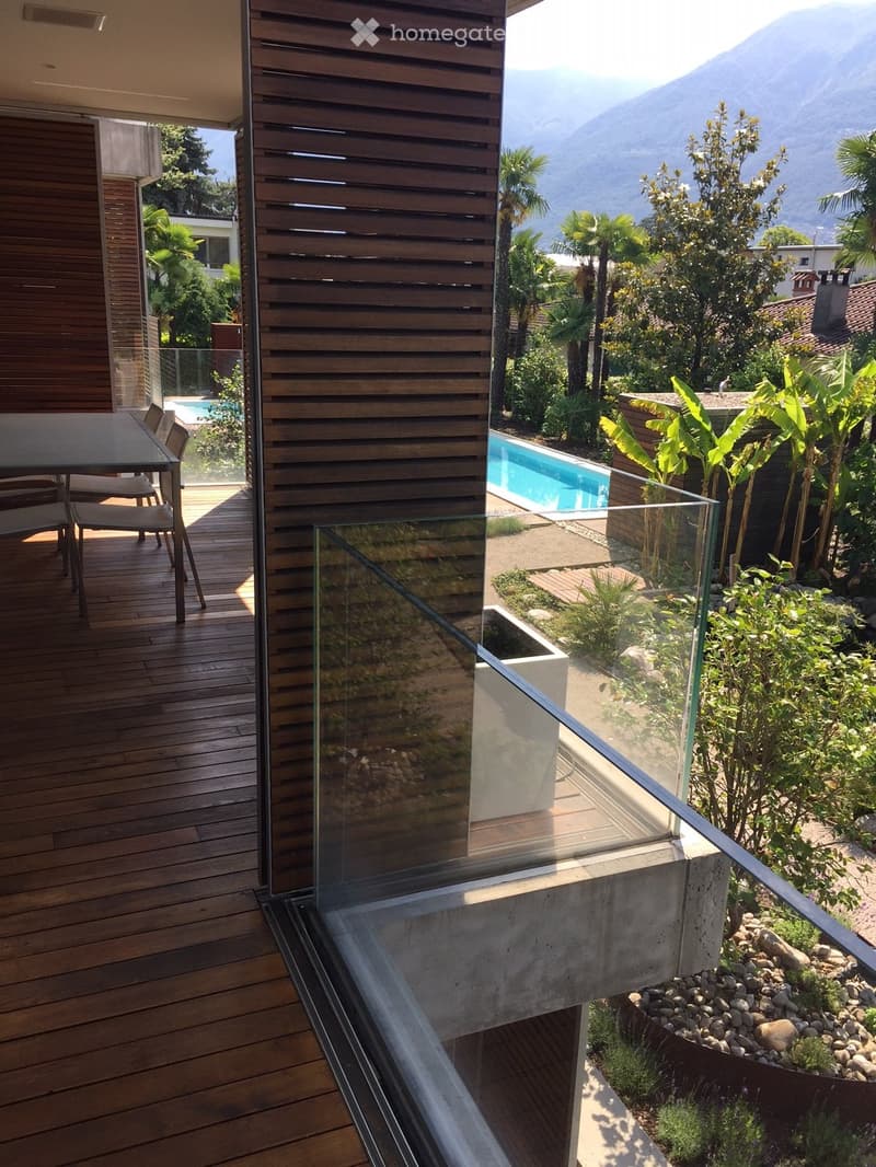 Balcony with wooden decking, glass railing, outdoor dining table and chairs, overlooking a landscaped garden with a swimming pool and mountain view.