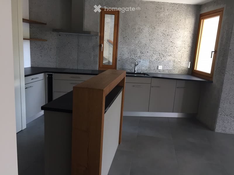Modern kitchen with grey cabinets, black countertops, a wooden island, and a sink with a faucet. The walls have a textured concrete finish, and there is a window on the right side.