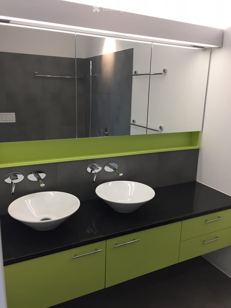 Modern bathroom with two white bowl sinks, sleek black countertop, green cabinets, and a large mirror with built-in lighting.