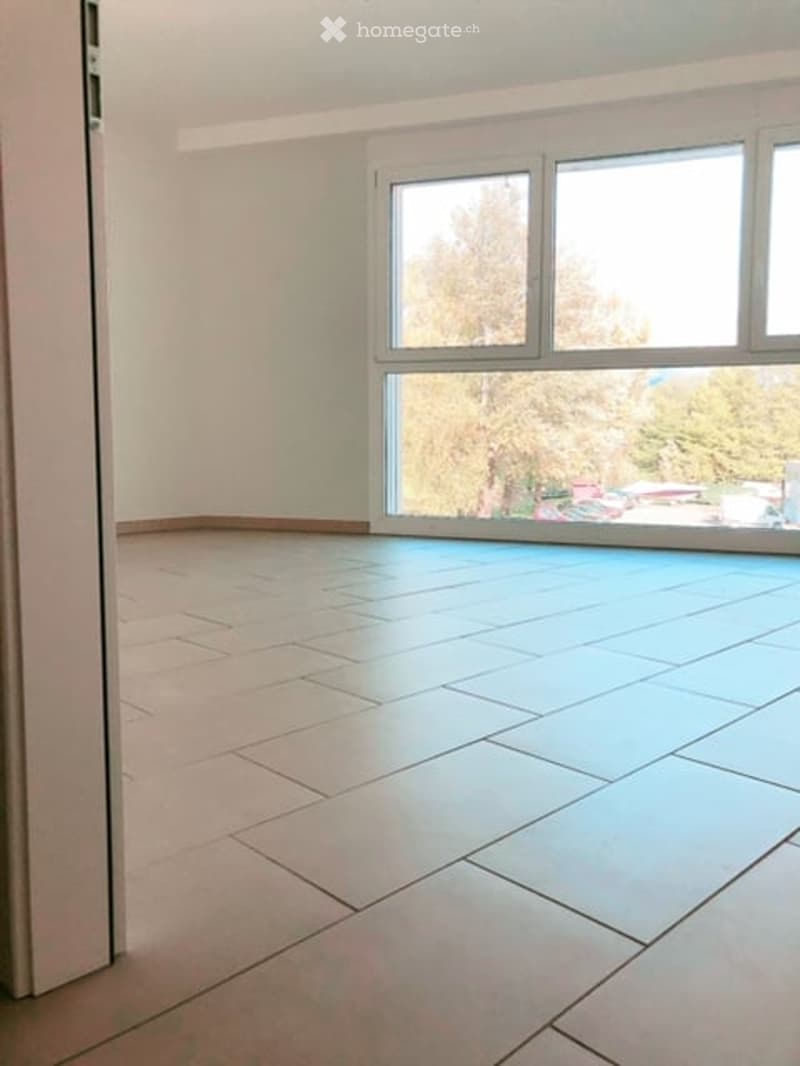 Empty room with white walls, tiled flooring, and large windows providing a view of trees outside.
