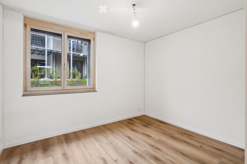 Empty room with light wooden flooring and white walls, featuring a large window with a view of a balcony and greenery outside.