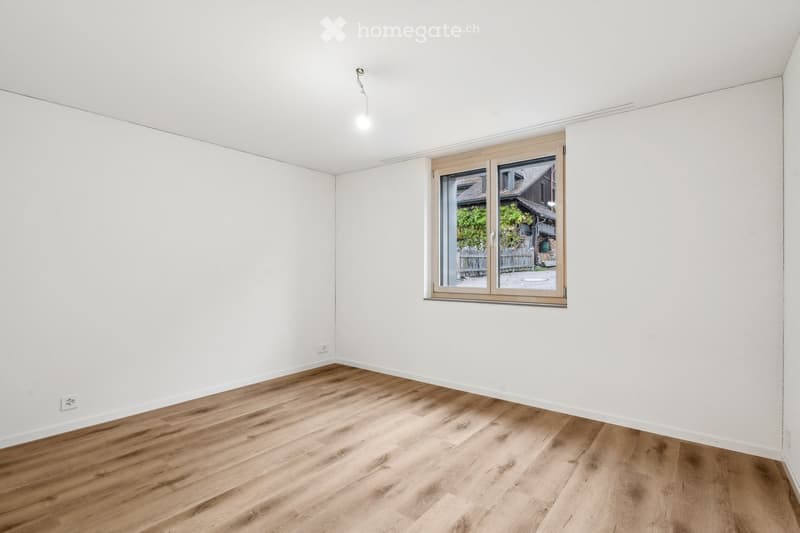 Empty room with white walls, wooden flooring, and a large window showing a view of a garden and house outside.
