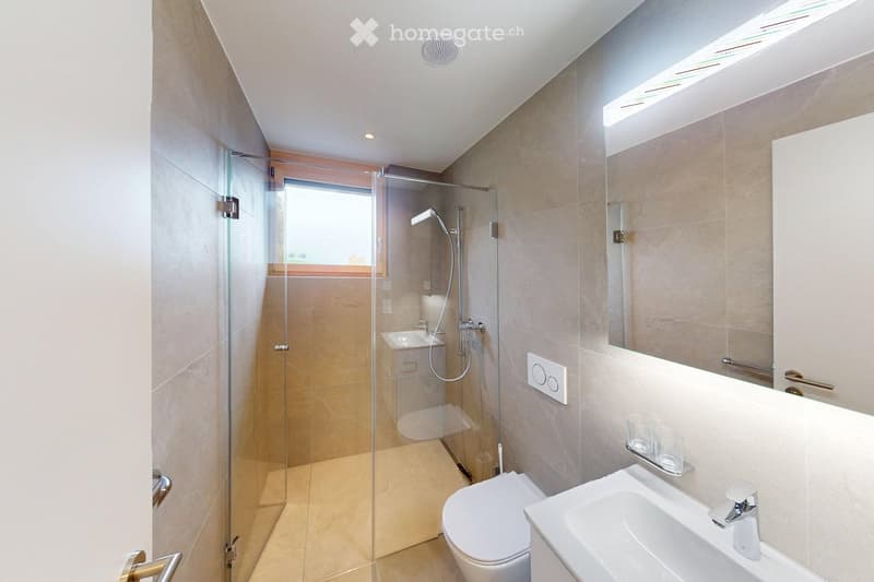 Modern bathroom with glass shower, white sink, white toilet, beige tiles, mirror, and a window