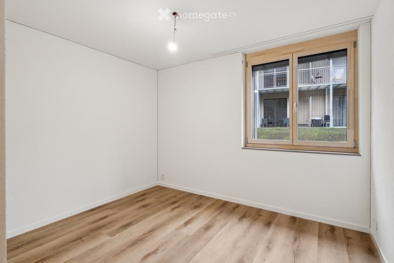 Empty room with light wooden flooring and white walls, featuring a large window with wooden frame and blinds, offering a view of an outdoor area with furniture.