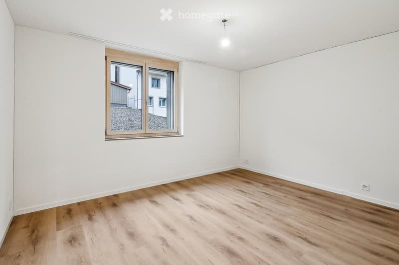 Empty room with light wooden flooring and white walls, featuring a large window with a view of the outside.