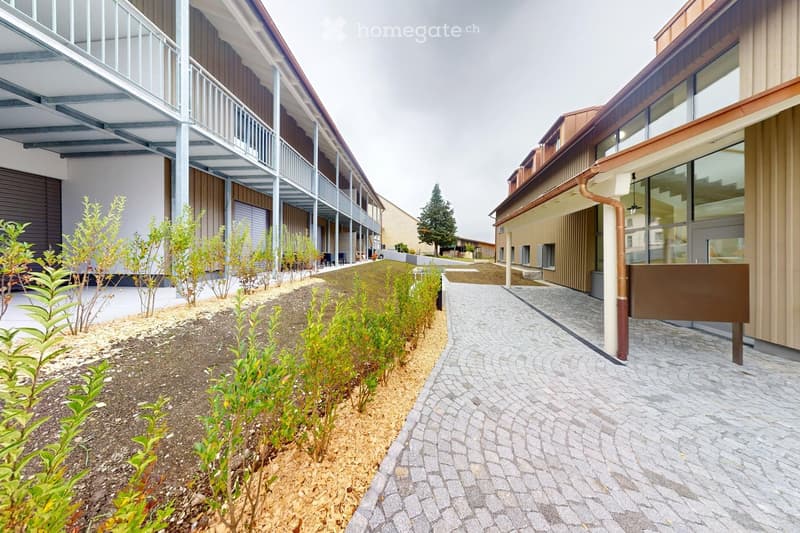 Two-story building with balconies, brick walls, glass windows, paved walkway, greenery