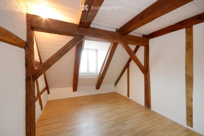 Empty attic room with exposed wooden beams, wooden floor, and a small window letting in natural light.