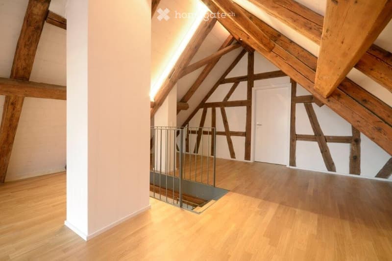 Empty attic space with wooden beams, exposed wooden ceiling, wooden floor, white walls, and a white door.