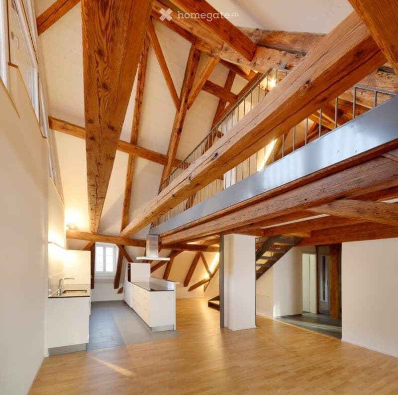 Open-plan space with exposed wooden beams, a modern white kitchen area, and a wooden staircase leading to an upper level.