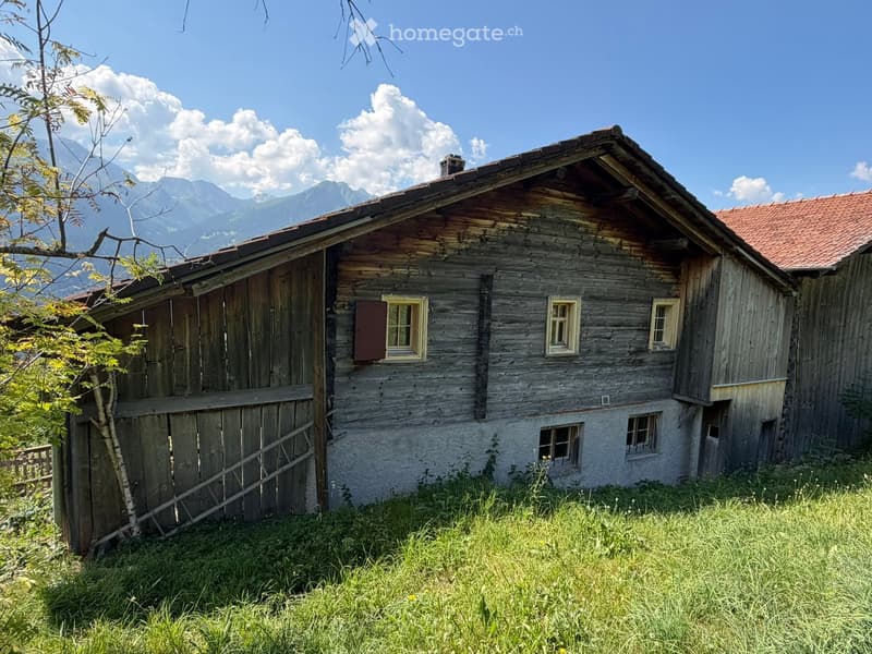 A rustic wooden house with a red-tiled roof, surrounded by grassy terrain and mountains in the background.