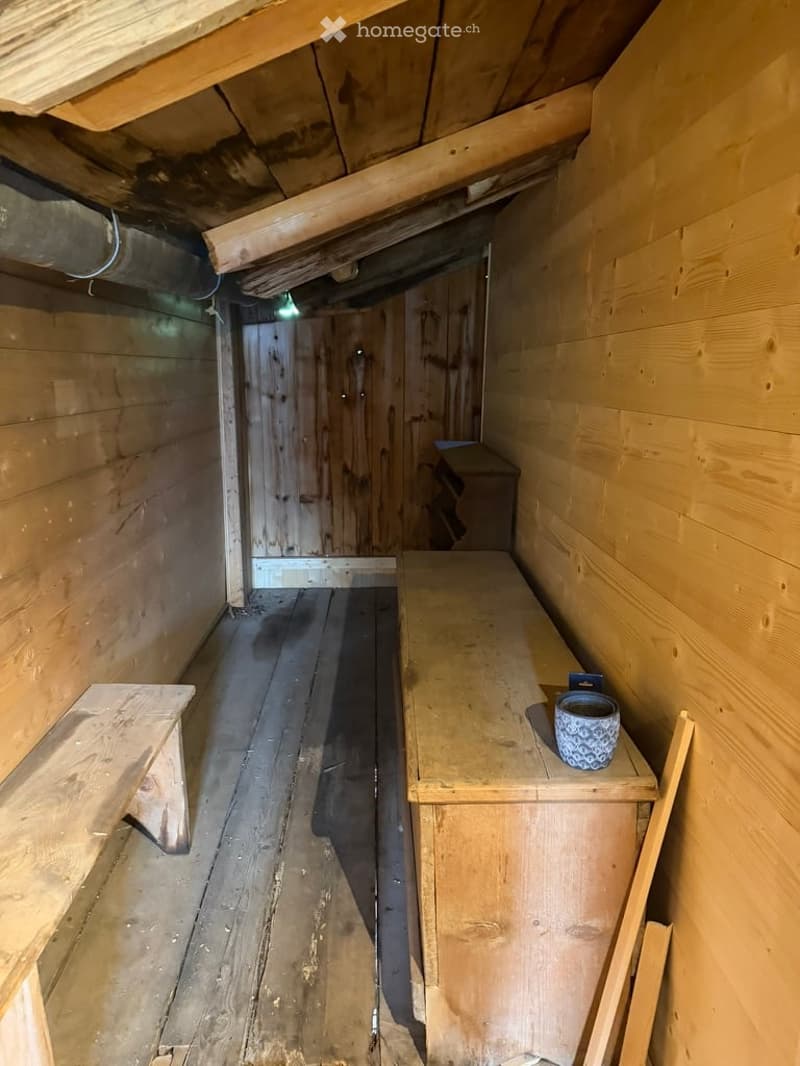A small, rustic wooden room with exposed wooden beams and a wooden floor. The room features a wooden bench, a small wooden table, and a wooden door leading to another space.