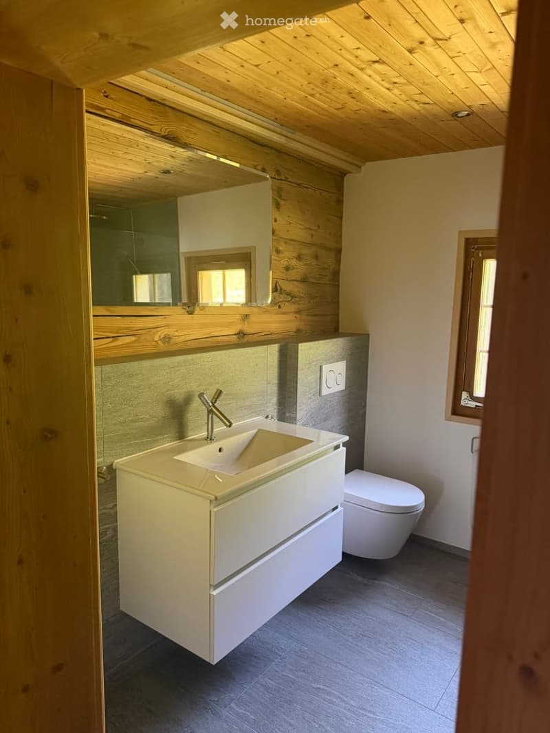 Modern bathroom with a white vanity, sleek sink, chrome faucet, and a large mirror above. The room features wooden accents and a grey tiled floor.
