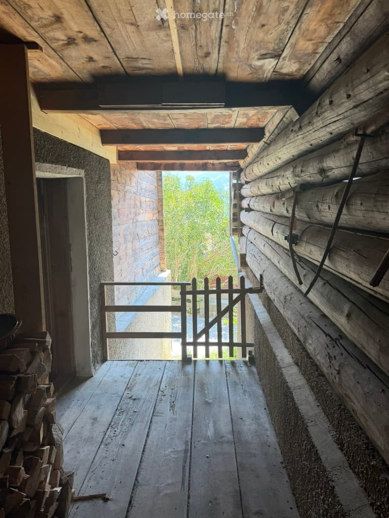 Wooden hallway with exposed beams, a metal gate leading outside, and a stack of firewood on the left side.