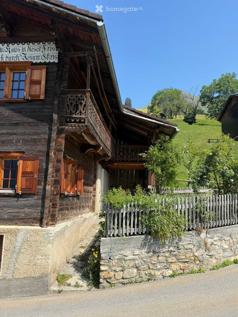 Traditional wooden house with wooden shutters, balcony, stone wall, and surrounding greenery.