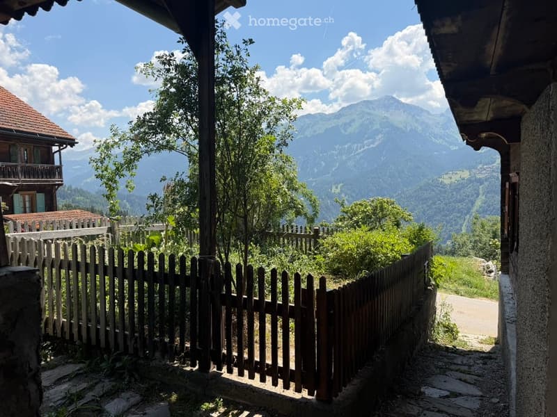 A wooden fence encloses a small garden area with lush greenery and a view of the surrounding mountains.