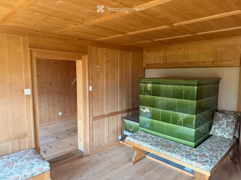 A small wooden room with a green tiled stove, two benches with floral upholstery, and wooden cabinets.