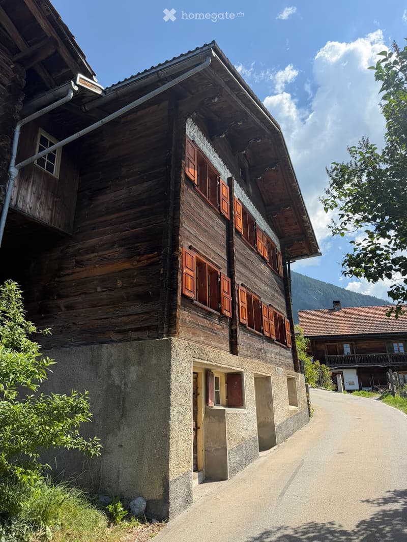 Traditional wooden house with multiple stories, wooden shutters, and a stone base, situated in a rural area with a clear view of mountains in the background.