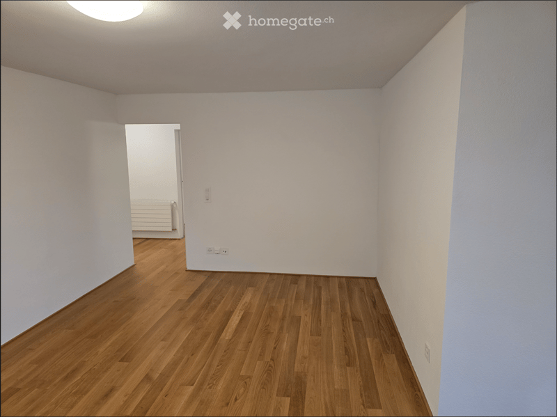 Empty room with wooden flooring and white walls, featuring a doorway leading to another room with a radiator visible.