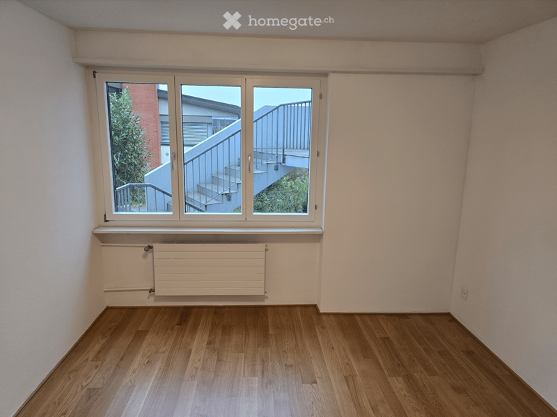 Empty room with wooden flooring, white walls, and a large window offering a view of a staircase and outdoor area.