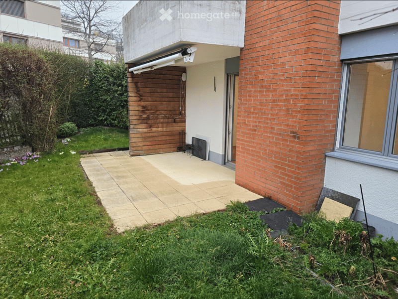 Garden area with a paved pathway leading to a wooden garage door, surrounded by grass and small plants. Adjacent to a brick building with a window and a small outdoor seating area.