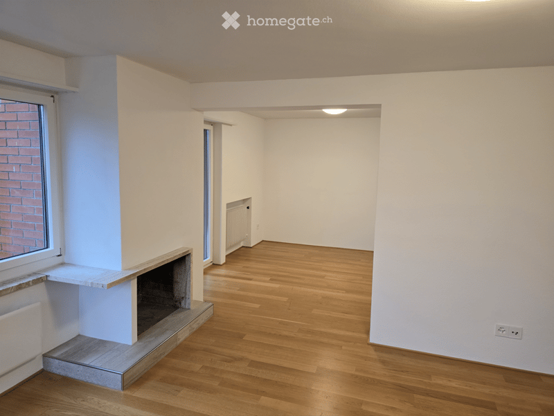 Empty room with wooden flooring, white walls, and a fireplace with a built-in shelf. There are two windows, one with a brick wall view, and a small hallway leading to another room.