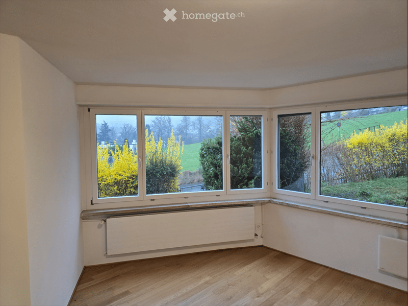 Empty room with wooden flooring, large windows offering a view of greenery, and radiators along the walls.