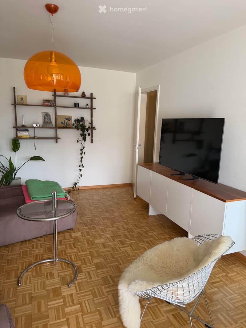 Modern living room with wooden herringbone flooring, a purple sofa, a round glass coffee table, a white wire chair with a fur throw, a large flat-screen TV on a wooden stand, and a wall-mounted shelf with books and decorative items.
