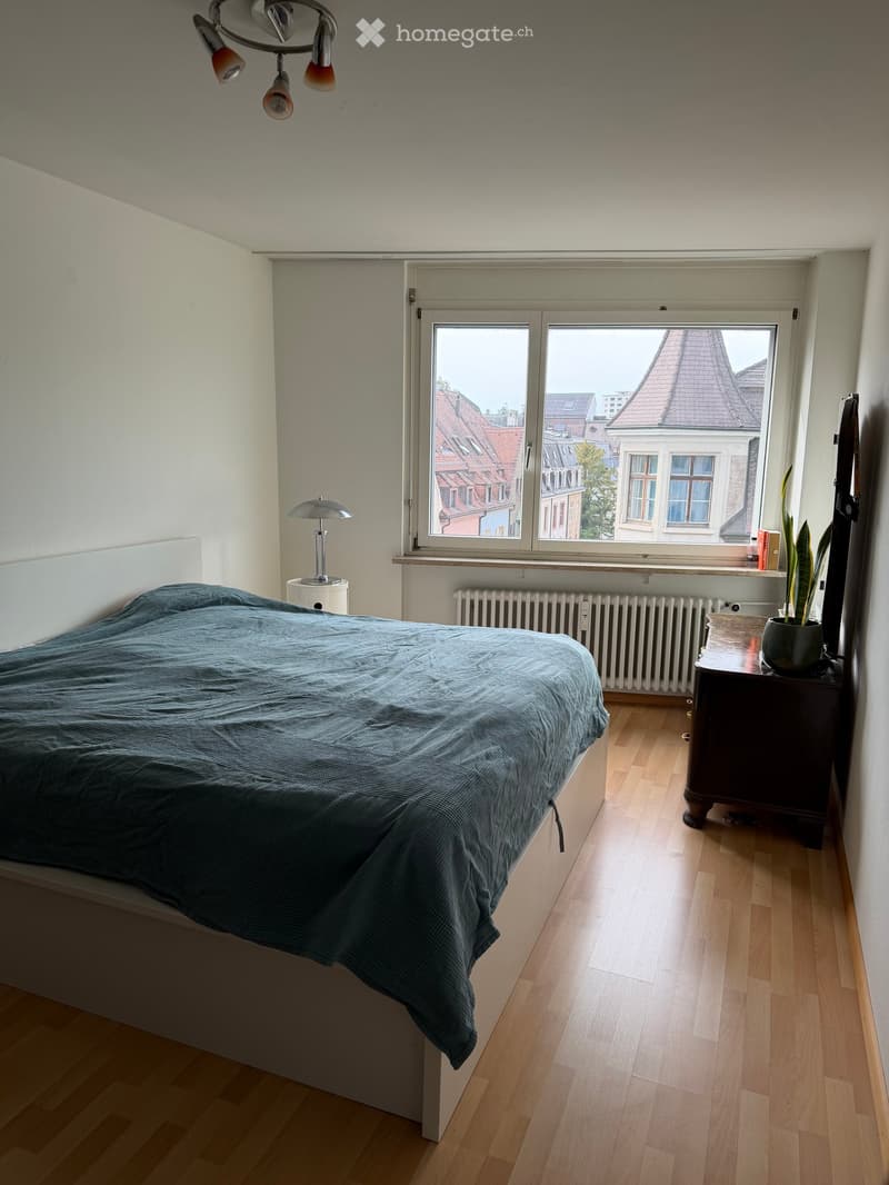 A modern bedroom with a wooden floor, a large window offering a cityscape view, a neatly made bed with a blue duvet, a nightstand with a lamp, and a potted plant on a small table.