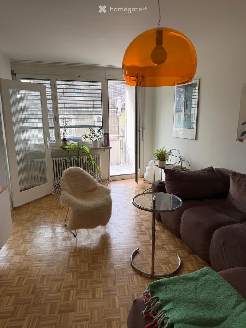 Modern living room with wooden herringbone flooring, large windows, a brown sofa, a glass coffee table, a white fur throw chair, and a hanging orange lamp.