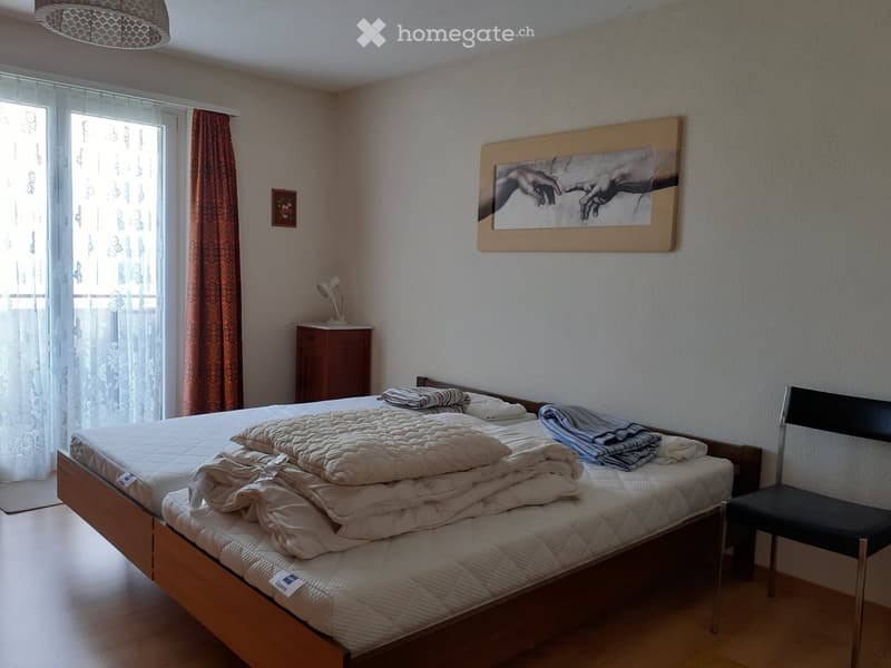 A well-lit bedroom with a double bed, neatly made with multiple blankets and pillows, a wooden nightstand with a lamp, a chair, and a large framed artwork on the wall.