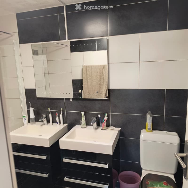 Modern bathroom with two white sinks, a mirrored cabinet, and a toilet. The walls are tiled in black and white, and various toiletries are placed on the sinks.