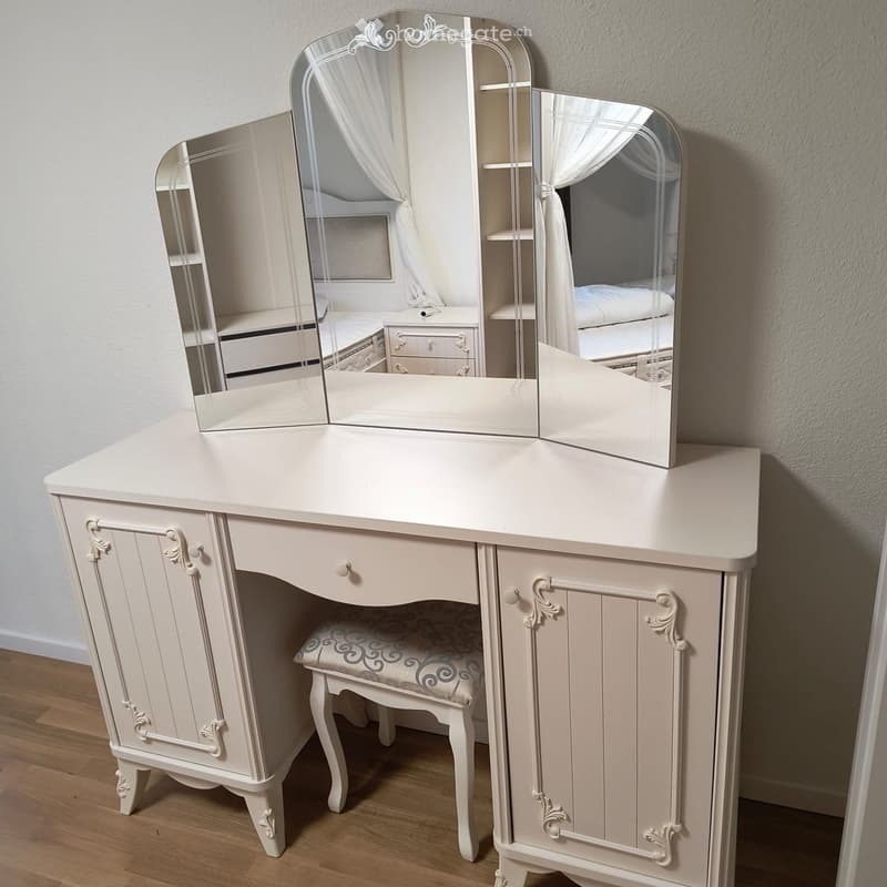 Elegant vanity with a three-panel mirror, ornate wooden frame, and a matching stool, reflecting a neatly made bed in the background.