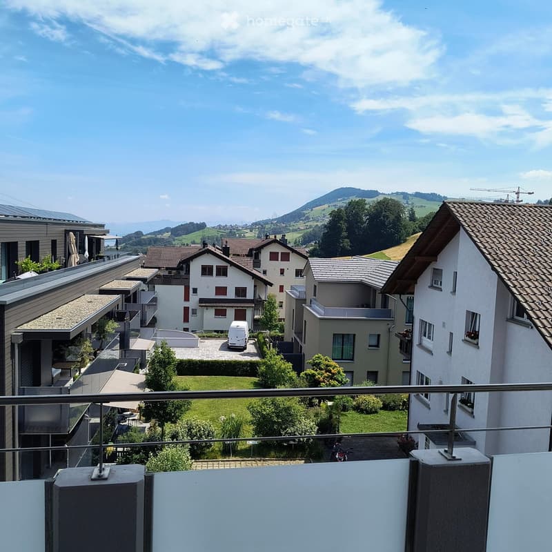 View from a balcony overlooking a residential area with houses, green lawns, trees, and distant hills under a clear sky.