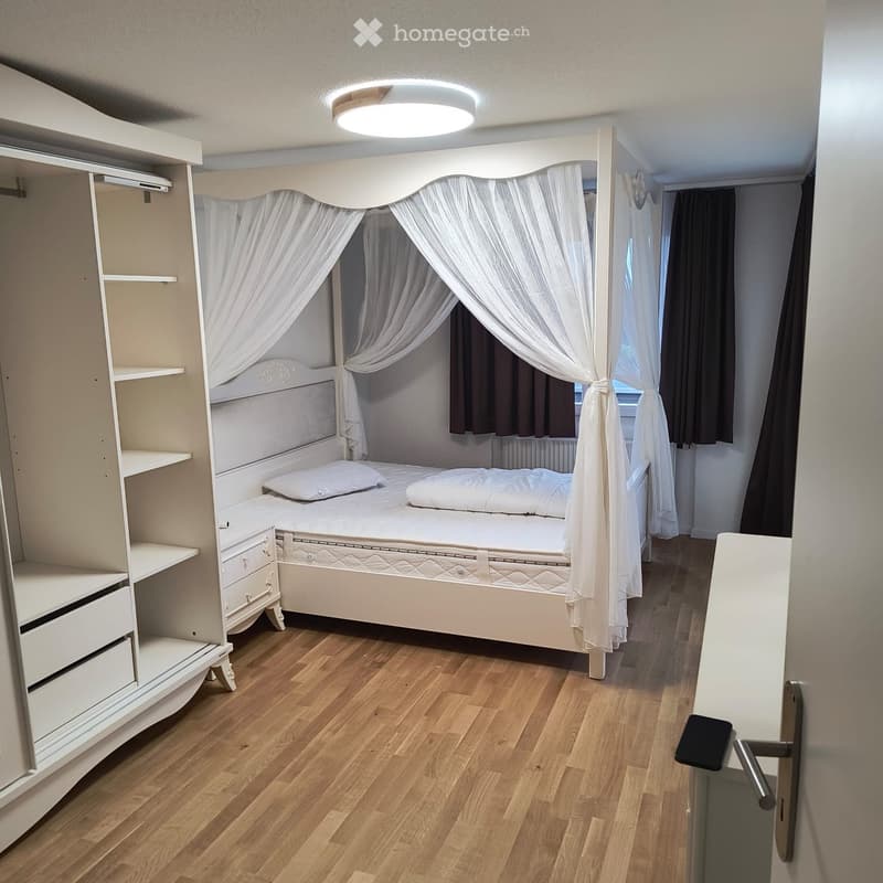 Cozy bedroom with a wooden floor, a large bed with a canopy, a bedside table, and a built-in wardrobe with shelves and drawers.