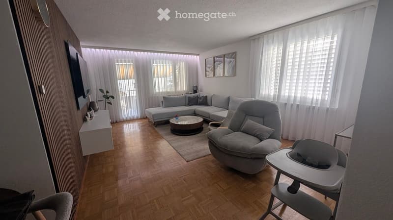 Modern living room with wooden flooring, large windows with white curtains, grey sectional sofa, coffee table, armchair, baby chair, small table, plant, and wall-mounted TV.