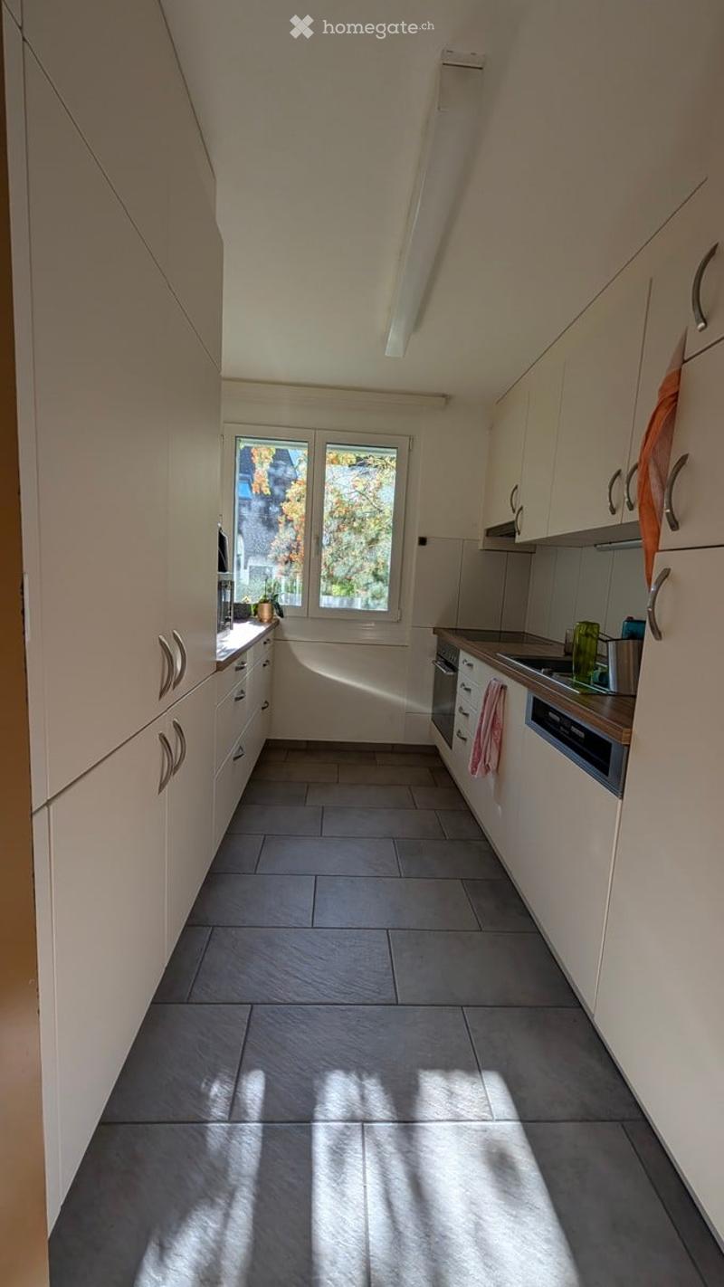 Modern kitchen with light-colored cabinets, dark tiled floor, and stainless steel appliances including an oven and dishwasher. Natural light streams in from a window above the sink.