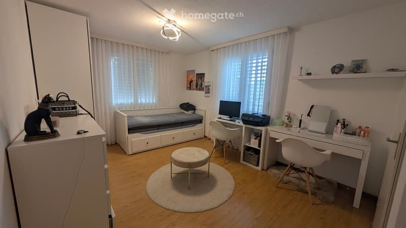 A well-lit room with a sofa bed, desk, chair, round stool, mirror, printer, cat statue, handbag, shelf, and picture frames.
