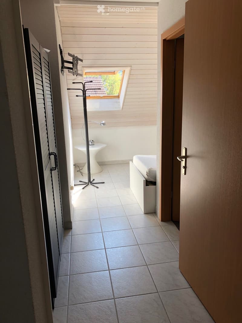 Small, furnished room with a bed, a white stool, a coat rack, and a lamp. The room has tiled flooring and a skylight window providing natural light.