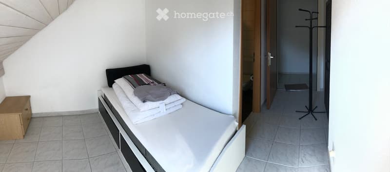 A small, modern bedroom with a single bed, nightstand, and neatly arranged bedding. The room features white walls, tiled flooring, and an open doorway leading to a hallway.